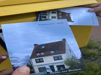 Eine Hand hält eine Postkarte, die ein Haus mit dem Aufdruck „Gruss aus Bremen“ zeigt, während eine andere Postkarte desselben Hauses in einen gelben Briefkasten eingeworfen wird.
