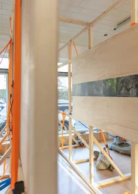 Holzrahmen mit großen Platten und orangefarbenen Bändern sind in einem Raum mit natürlichem Licht aufgestellt. Auf dem Boden sind Felsen und Kabel zu sehen, und die Fenster zeigen Autos und Grünflächen im Freien.