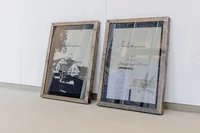 Zwei gerahmte Dokumente mit Holzumrandung liegen auf dem Boden und lehnen an einer weiß strukturierten Wand. Auf dem einen Rahmen ist ein Gebäude abgebildet, auf dem anderen ein Text und die Skizze eines Schlüssels.