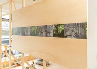 Auf einer großen Holztafel ist eine Serie von vier Fotografien zu sehen, die Nahaufnahmen einer von Grün umgebenen Steinskulptur zeigen, die in einem Innenraum aufgestellt ist und in deren Nähe Baumaterialien zu sehen sind.