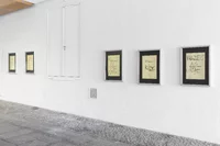 Fünf gerahmte Poster mit handgeschriebenem Text und Skizzen hängen an einer weißen Wand entlang eines Kopfsteinpflasterwegs in einem hellen, minimalistischen Galerieraum. Die Poster enthalten Sätze wie „Schlüssel zur Zukunft“ und „Business as usual“.