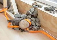 Auf einem hellen Holzboden neben einer Holzkonstruktion und einer Fensterbank liegt ein Stapel Steine unterschiedlicher Größe, der von einem orangefarbenen Band zusammengehalten wird. Einige Steine sind auf der Fensterbank verstreut.