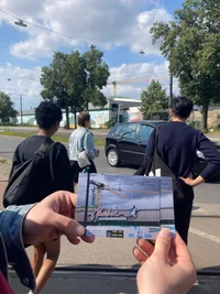 Eine Person hält eine Postkarte mit Straßenkunst, während drei Personen an einem sonnigen Tag an einem Zebrastreifen mit vorbeifahrenden Autos, Bäumen und Gebäuden im Hintergrund stehen.