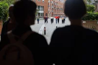 Zwei Personen in Silhouette beobachten eine Gruppe von Menschen in bunten Jacken, die mit ausgestreckten Armen in der Ferne auf einer Kopfsteinpflasterstraße zwischen Backsteinwohnhäusern stehen.