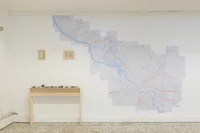 Eine große Landkarte ist an einer weißen Wand über einem einfachen Holztisch angebracht, auf dem Steine oder kleine Gegenstände angeordnet sind. Links neben der Karte hängen zwei kleine gerahmte Bilder.
