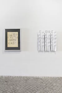 Ein gerahmtes Papier mit dem handgeschriebenen Text CENSORED FORGOTTEN HIDDEN MEMORY (ZENSIERTE VERGESSENE VERGESSENE ERINNERUNG) hängt an einer weißen Wand neben einer Auslage mit mehreren weißen Flugblättern, die in Reihen angeordnet sind. Der Boden besteht aus gemusterten Steinfliesen.