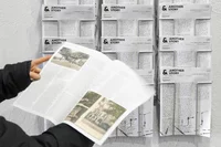 Eine Person hält ein aufgeschlagenes Magazin mit Fotos und Text in der Hand, während an der Wand mehrere Exemplare einer Publikation mit dem Titel Another Story vor einer weißen Wand ausgestellt sind.