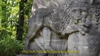 Eine verwitterte steinerne Löwenskulptur steht in einem von Grün umgebenen Park, mit gelben Untertiteln am unteren Rand, die lauten: (der Löwe hat Schwierigkeiten beim Atmen).