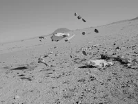 Knochen und Steine schweben unter klarem Himmel über einer kargen, felsigen Wüstenlandschaft. Im Hintergrund sind Berge zu sehen. Das Bild ist schwarz-weiß.