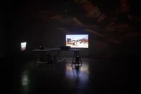 Ein schwach beleuchteter Raum mit einem großen Tisch in der Mitte, auf dem zwei Projektoren stehen. An der Wand hinter dem Tisch sind zwei rechteckige Projektionen von Außenszenen zu sehen. Der Boden glänzt und reflektiert etwas Licht.