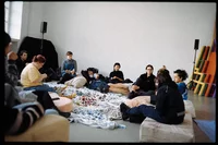 Eine Gruppe von Menschen sitzt in einem hellen Raum auf Kissen und Decken und unterhält sich. Künstlerbedarf und bunte Materialien liegen verstreut auf dem Boden, im Hintergrund lehnen bunte Gegenstände an der Wand.