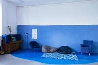 Ein gemütlicher Raum mit blauen Wänden und blauem Boden, einem gemusterten Teppich, zwei Sitzsäcken, einem blauen Sessel, einem Klavier mit einer Blumenvase, einer Stehlampe und einem blauen Polsterstuhl. An der Wand hängen Poster.