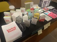 Ein Ausstellungstisch mit verschiedenen kleinen Broschüren, Zines und Papierstapeln mit dem Titel „Living Room“, daneben eine Kaffeetasse und handgeschriebene Haftnotizen mit Preisen und Etiketten.