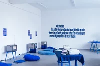 Eine Galerie für zeitgenössische Kunst mit weißen Wänden, blauen Möbeln, niedrigen Tischen, Sitzkissen, drei kleinen Fernsehern und blauer Textkunst an der Wand. Broschüren und Bücher liegen auf Tischen. Der Raum ist hell erleuchtet.