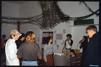 Eine Gruppe von Menschen trifft sich in einem geschlossenen Raum. Zwei Personen singen in Mikrofone. Kunstwerke schmücken die Wände, und darüber hängt ein Rauchverbotsschild. An der Decke hängen verdrillte Drähte oder Dekorationen.