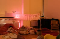 In einem schwach beleuchteten Raum mit warmer Beleuchtung entspannen sich Menschen auf Sitzsäcken und Decken. Zwischen den Garderobenständern hängt ein Spitzenbanner mit Text. Im Hintergrund stehen ein gepolsterter Sessel und ein Heizkörper.