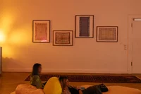 Zwei Personen sitzen auf weichen Sitzgelegenheiten in einem warm beleuchteten Raum mit beigen Wänden. Über ihnen hängen vier gerahmte Textilkunstwerke, und auf dem Holzboden neben einer geschlossenen weißen Tür liegt ein gemusterter Teppich.