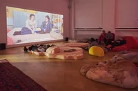 Menschen sitzen und liegen auf großen Kissen und Sitzsäcken in einem schwach beleuchteten Raum und schauen sich einen Film an der Wand an. Auf dem Bildschirm sind zwei Personen zu sehen, die sich in gemütlicher Atmosphäre unterhalten; Untertitel sind zu sehen.