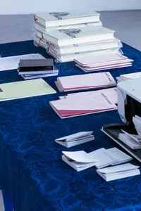 Eine blaue Tischdecke bedeckt einen Tisch, auf dem Stapel von Büchern, rosa und grünen Papieren, Broschüren und Dokumenten liegen. Am Rand ist ein Teil eines Druckers zu sehen.