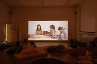 Ein schwach beleuchteter Raum mit Menschen, die auf Bodenkissen sitzen und liegen und einen an die Wand projizierten Film ansehen. Der Film zeigt drei Frauen, die zusammensitzen. Englische Untertitel sind zu sehen.