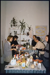 Sechs Personen versammeln sich um eine Kücheninsel, die mit Essen, Getränken und Kochzutaten bedeckt ist. Regale mit Pflanzen und Tassen schmücken die Wand dahinter. Auf einem gelben Plakat steht: „HOW (NOT) TO BUILD A...”. Sie scheinen sich zu unterhalten.