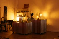 Ein warm beleuchtetes Café mit einer Person hinter einem Tresen, umgeben von Regalen voller Waren. Das Ambiente ist gemütlich, sanftes gelbes Licht und ein handgeschriebenes „Living Room“-Schild über den Regalen.