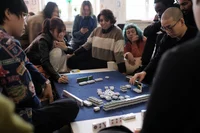 Eine Gruppe von Menschen sitzt um einen mit einem dunklen Tuch bedeckten Tisch und spielt ein Spiel mit Spielsteinen, möglicherweise Mahjong. Einige konzentrieren sich auf das Spiel, andere schauen zu oder benutzen ihre Handys. Der Raum wirkt hell und gemütlich.