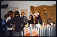 Eine Gruppe von Menschen versammelt sich um eine kleine Bar oder einen Kiosk mit Getränken, Speisekarten und Blumen auf der Theke. Drei Personen stehen hinter der Theke, zwei Personen stehen davor und unterhalten sich.