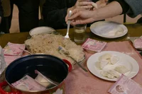 An einem mit Tellern, einem Topf mit kochendem Wasser, Teigtaschen, Instant-Nudelpackungen und einem Glas Wasser gedeckten Tisch bereiten Menschen Teigtaschen und Nudeln zu; Hände formen Teigtaschen und servieren das Essen.