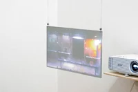 Ein Projektor auf einem Holzregal projiziert ein buntes, verschwommenes Bild auf eine transparente Leinwand, die an Drähten in der Ecke eines weißen Raums aufgehängt ist.