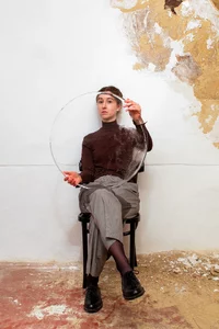 Eine Person sitzt auf einem Stuhl an einer abgenutzten Wand und hält ein großes, durchscheinendes, rundes Objekt vor ihren Oberkörper und ihr Gesicht, wodurch eine verzerrte Spiegelung entsteht. Boden und Wand zeigen abblätternde Farbe und Schutt.