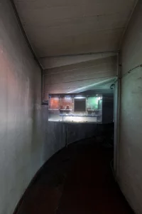 Ein schwach beleuchteter, geschwungener Betonflur mit fleckigen Wänden. Zentral hängt eine kleine LEinwand mit einer Videoprojektion welche die Rückseite einer beleuchteten Bushaltestelle zeigt.