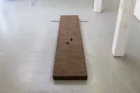 Eine lange, rechteckige braune Platte aus Ton liegt flach auf einem hellgrauen Boden in einem minimalistischen, industriell gestalteten Raum. An den Ecken sind Metallstangen und Spannelemente angebracht, und auf der Platte sind zwei kleine quadratische Aussparungen sichtbar.