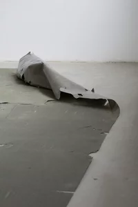 Ein Stück grauen, zerrissenen Materials liegt teilweise aufgerollt auf einem glatten, hellgrauen Boden in einem minimalistischen, leeren Raum mit weißen Wänden.