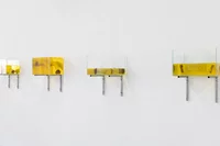 Vier rechteckige Glastanks, gefüllt mit gelber Flüssigkeit (Öl) und jeweils einem schwimmenden Gegenstand, sind in einer Reihe auf Metallhalterungen an einer weißen Wand montiert.