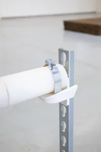 Eine weißes zylindrischen Stange ist mit einer Metallklemme und einer weißen Halterung an einem vertikalen Metallstützrahmen befestigt, der auf einem glatten grauen Boden liegt.