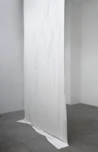 Ein hoher, schmaler weißer Stoff oder ein Laken hängt vertikal von der Decke bis zum Boden in einem minimalistischen, leeren Raum mit grauem Betonboden und schlichten weißen Wänden.