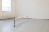 Ein leerer, minimalistischer Raum mit einem großen Fenster und einer weißen zylindrischen Stange, die auf Metallstützen auf dem schlichten, hellgrauen Boden steht.