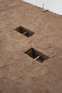 Zwei quadratische Löcher in einer strukturierten Oberfläche aus braunem Ton, jedes mit einem Metallscharnier oder einer Halterung, wobei an der oberen rechten Kante eine weitere Metallbefestigung sichtbar ist.