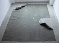 Ein großes, graues, zerknittertes Laken liegt ausgebreitet auf dem nackten Boden in einem schlichten weißen Raum, wobei zwei Ecken des Lakens teilweise nach oben gebogen sind.