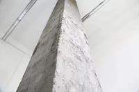 Nahaufnahme, Untersicht einer hohen Betonsäule mit rauer Struktur, die sich nach oben zu einer weißen Decke mit sichtbaren Rohren oder Leitungen erstreckt.