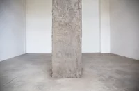 Eine große Betonsäule steht in der Mitte eines leeren, unfertigen Raums mit schlichten weißen Wänden und einem nackten Betonboden.