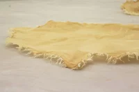 Ein Stück hellbrauner, ausgefranster Stoff liegt flach auf einer blassen Oberfläche, mit gezackten Kanten und ungleichmäßiger Textur. Ein weiterer ähnlicher Stoff ist unscharf im Hintergrund zu sehen.