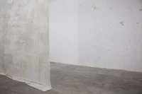 Ein minimalistischer, leerer Raum mit Betonboden und schlichten weißen Wänden. Ein transparenter, cremefarbener Stoff oder Vorhang hängt von der Decke und unterteilt den Raum teilweise. Die Wände weisen schwache Flecken und Flecken auf.