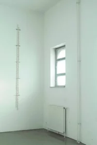Ein minimalistischer, lichtdurchfluteter weißer Raum mit einem kleinen Fenster, einem Heizkörper darunter, freiliegenden vertikalen Rohren und einer vertikalen Reihe weißer, ovaler Objekte, und Metallverbindungen, die an der linken Wand befestigt sind.