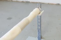 Eine Nahaufnahme einer weißen zylindrischen Stange, das mit einer Klammer an einer vertikalen Metallhalterung befestigt ist, vor dem Hintergrund eines grauen Betonbodens.