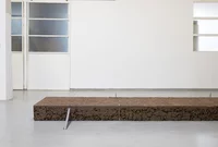 Eine rechteckige Platte aus braunem Ton ruht auf einem Galerieboden mit weißen Wänden und großen Türen; unter der Platte sind eine Metalstange und Spannelemente angebracht.