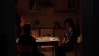 Zwei Personen sitzen sich an einem schwach beleuchteten Küchentisch gegenüber, wobei das warme Licht Schatten wirft. Im Hintergrund sind Küchenregale, ein Herd und Schränke zu sehen.