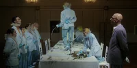 Mehrere Personen in hellblauen Anzügen und Handschuhen stehen oder knien um einen langen Esstisch; eine Person steht auf dem Tisch und schüttet Flüssigkeit aus, während eine andere kniet und eine Sauerei aufräumt. Die Szene wirkt inszeniert und dramatisch.