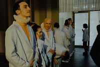 Eine Reihe von Personen in blassblauen Anzügen, mit Nackenbügeln und blauen Handschuhen, steht in einem Innenraum. Einige blicken nach vorne, andere nach unten; zwei Personen unterhalten sich im Hintergrund in der Nähe eines hellen Fensters.